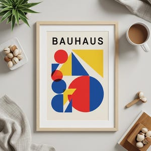 Pode incluir: Impressão de arte Bauhaus emoldurada com um design geométrico em vermelho, azul e amarelo. A palavra "BAUHAUS" é impressa em preto no topo. A impressão está em uma moldura de madeira clara, em um fundo branco.