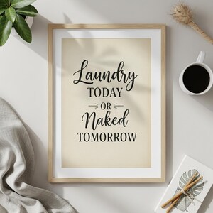 Può includere: Una stampa incorniciata con il testo "Laundry Today or Naked Tomorrow" in scrittura nera. La stampa ha uno sfondo beige chiaro e una cornice in legno naturale. Sono visibili anche una piccola pianta, una tazza di caffè e un oggetto decorativo.