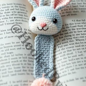 May include: A crocheted rabbit bookmark in blue, white, and pink with a pink pom-pom at the bottom. The bookmark is on top of an open book.
