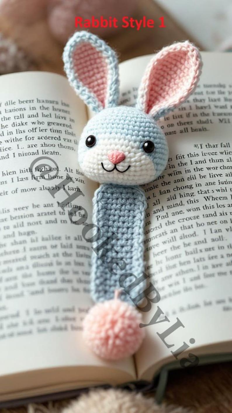 May include: A crocheted rabbit bookmark in blue, white, and pink with a pink pom-pom at the bottom. The bookmark is on top of an open book.