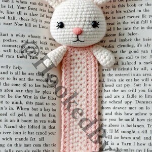 May include: A white crocheted rabbit bookmark with pink ears and a pink pom-pom at the bottom. The bookmark is on a book with text.