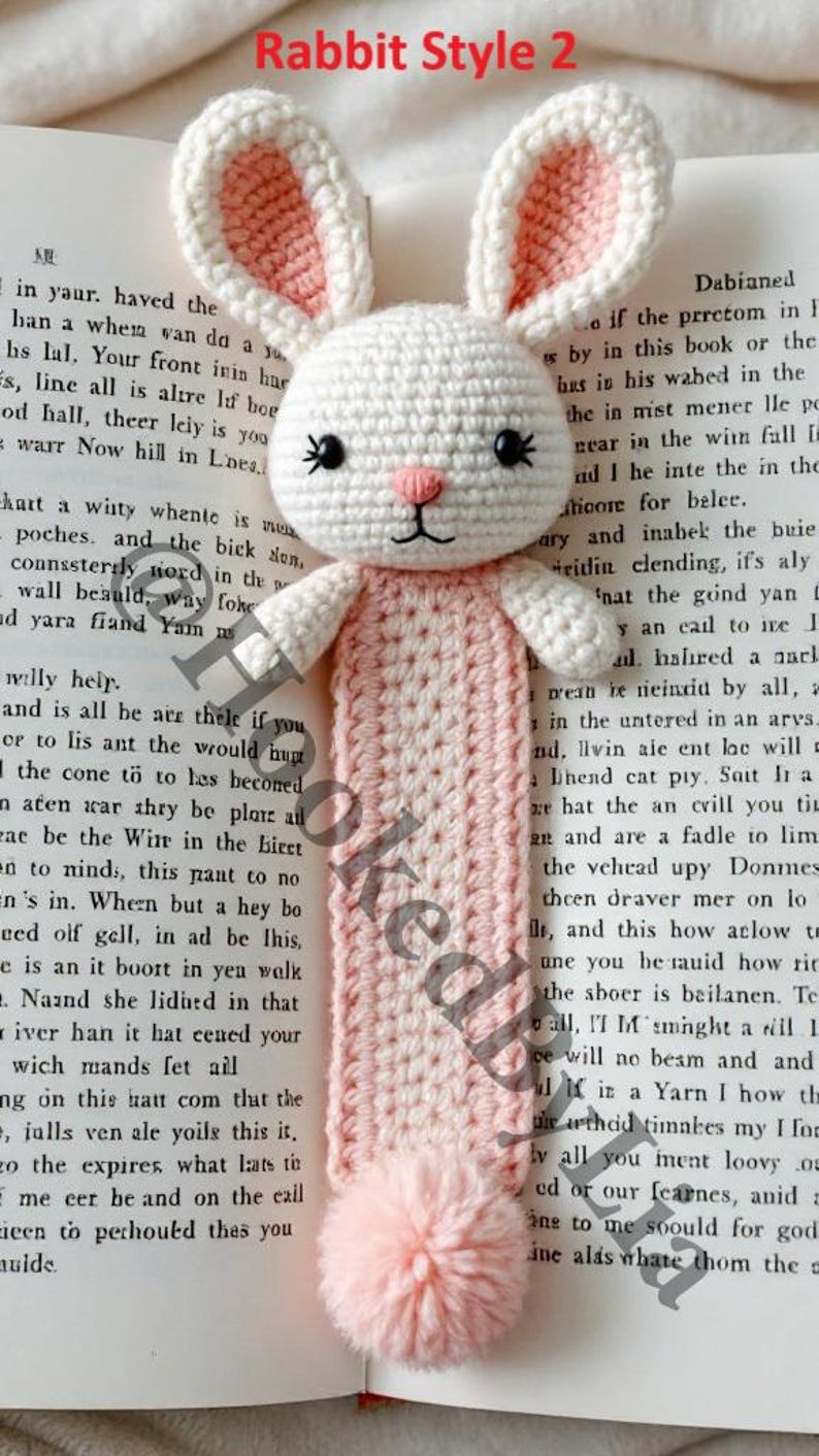 May include: A white crocheted rabbit bookmark with pink ears and a pink pom-pom at the bottom. The bookmark is on a book with text.