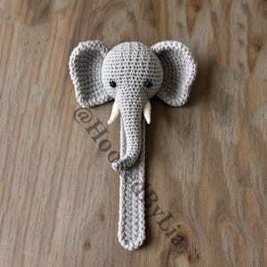 Op de afbeelding: Een grijs gehaakt olifantenspeelgoed met een lange slurf en grote oren. De olifant heeft zwarte ogen en een wit gehaakt slagtand aan elke kant van zijn slurf.