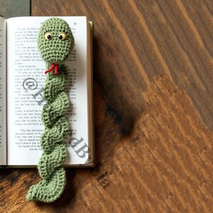 May include: Green crocheted snake bookmark with large eyes and a red tongue. The bookmark is laying on a book.