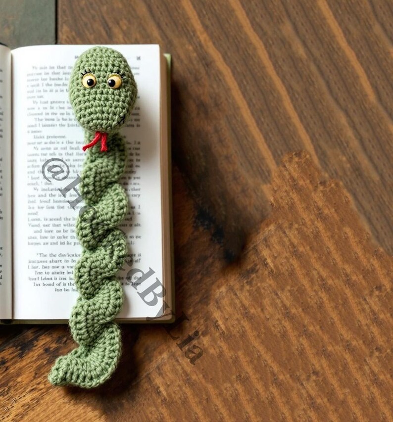 May include: Green crocheted snake bookmark with large eyes and a red tongue. The bookmark is laying on a book.