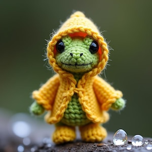 Könnte beinhalten: Eine gehäkelte Froschfigur mit gelber Kapuzenjacke und passenden Stiefeln. Der Frosch ist grün mit großen schwarzen Augen und einem lächelnden Gesichtsausdruck. Die Jacke hat eine geflochtene Bordüre und Knopfverschlüsse. Der Hintergrund ist verschwommen grün.