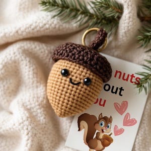 May include: A crocheted acorn keychain with a smiling face and brown cap. The keychain is hanging on a white fuzzy surface with a card that says "nuts about you" with hearts.