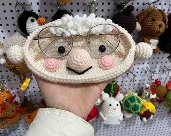 May include: A handmade, off-white crochet eyeglass holder shaped like a face, complete with round glasses, a pink nose and cheeks, and black eyes. Other crochet keychains are visible in the background.