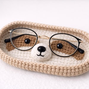 Peut inclure: Un plateau ovale en crochet beige présentant un museau central blanc avec un nez noir et des taches marron. Une paire de lunettes à monture noire avec des branches dorées repose sur le plateau, positionnée sur les taches marron.