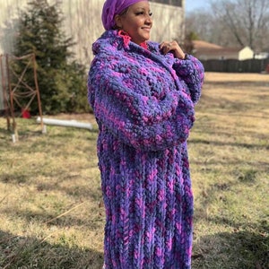 May include: A long, hooded robe in shades of purple and pink. The robe is made of a thick, textured material, and the hood is up. The person in the image is wearing a matching purple head covering.