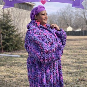 May include: A woman wearing an oversized, long crochet sweater in shades of purple and pink. The sweater has a hood and is made of thick yarn. The image includes text that reads "CROCHET PATTERN Oversized Long Sweater" and "Instant Download PDF Pattern".