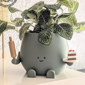 Peut inclure: Un pot en céramique vert en forme de personnage de dessin animé avec un visage souriant, tenant un rouleau à pâtisserie d'une main et une assiette avec une part de gâteau de l'autre. Le pot a de petits pieds et est rempli d'une plante verte feuillue.