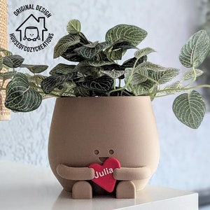 Könnte beinhalten: Ein brauner Keramiktopf in Form einer Person, die ein rotes Herz mit dem Namen "Julia" hält. Der Topf hat ein kleines Lächeln und zwei Augen. Eine grüne Pflanze mit weißen Adern wächst im Topf.