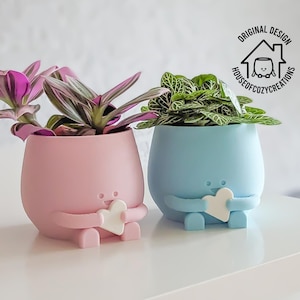 Op de afbeelding: Twee kleine, kleurrijke, 3D-geprinte potten met schattige gezichtjes en hartjes. De potten bevatten kleine plantjes. De roze pot heeft een paarse en groene plant, en de blauwe pot heeft een groene plant.