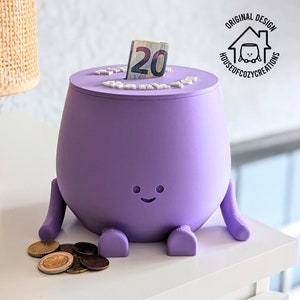 Peut inclure: Tirelire violette avec un visage souriant, des bras et des jambes. Un billet de vingt euros est inséré dans la fente sur le dessus de la tirelire. La tirelire est posée sur une surface blanche.