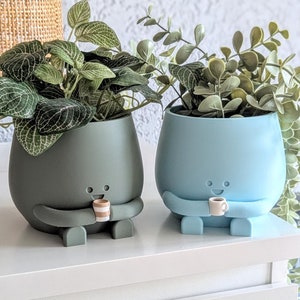 May include: Two ceramic plant pots with cartoon faces and arms holding small cups. The pot on the left is dark green and the pot on the right is light blue. Both pots have plants growing out of them.