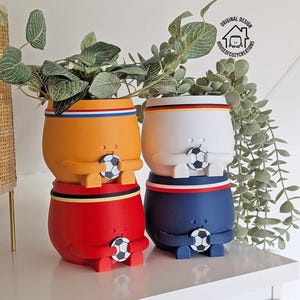 Op de afbeelding: Vier ronde plantenbakken in de vorm van figuren die voetballen vasthouden. De plantenbakken zijn oranje, wit, rood en marineblauw, elk met een gekleurde rand en een kleine voetbal in het midden. Een logo met de tekst ORIGINAL DESIGN HOUSEOFCOZYCREATIONS is zichtbaar.