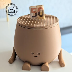 May include: A brown ceramic money box with a smiling face and two small legs. The money box has a waffle-patterned lid and a 50 Euro note is inserted into the slot.