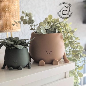 Può includere: Due vasi in ceramica con facce sorridenti e gambe. Il vaso a sinistra è verde e contiene una succulenta. Il vaso a destra è marrone e contiene una pianta a foglia. I vasi sono su una superficie bianca.