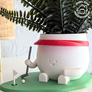 Op de afbeelding: Een witte keramische plantenpot in de vorm van een golfer met een rode hoed, die een golfclub vasthoudt en op een groene golfbaan staat. De pot heeft een vrolijk gezicht en houdt een golfclub vast. De pot staat op een wit oppervlak.