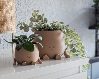 May include: Two brown ceramic planters with smiling faces and legs, holding green plants. The planters are on a white surface.