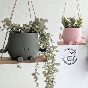 May include: Two hanging planters, one grey and one pink, each with a smiling face and arms and legs, resembling a character. The planters are suspended by twine and hold greenery. The planters sit on a wooden swing.