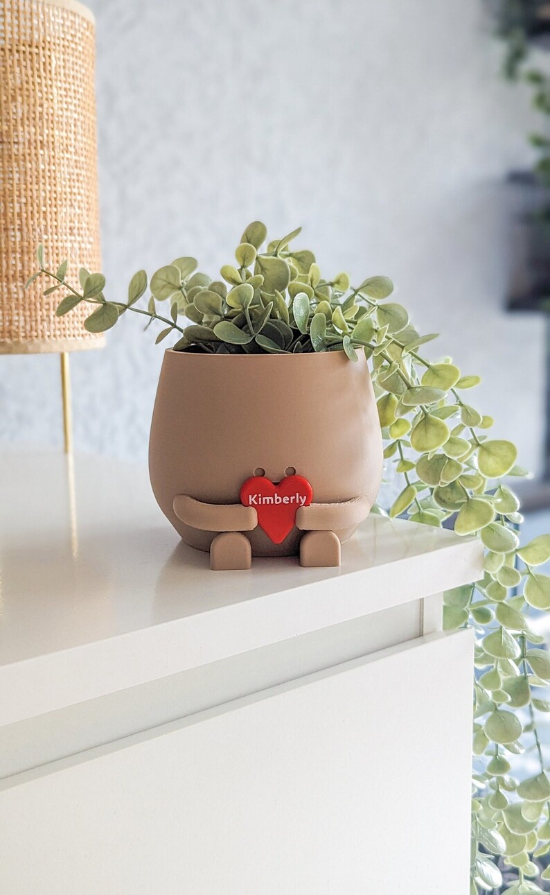 Puede incluir: Un macetero de cer&aacute;mica marr&oacute;n con forma de persona que sostiene un coraz&oacute;n rojo con el nombre "Kimberly" en &eacute;l. El macetero est&aacute; lleno de una planta verde de hojas.