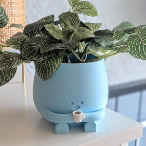 May include: A light blue ceramic planter shaped like a person holding a small white cup. The planter has a happy face and is sitting on a white surface. The planter is filled with a green leafy plant.
