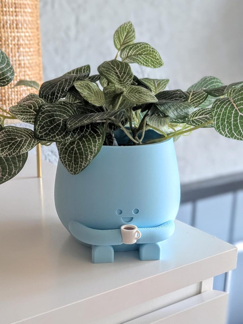 May include: A light blue ceramic planter shaped like a person holding a small white cup. The planter has a happy face and is sitting on a white surface. The planter is filled with a green leafy plant.
