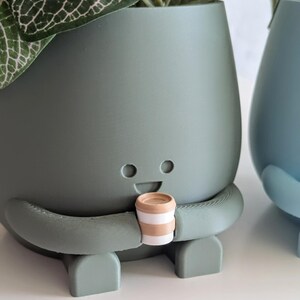 May include: A green ceramic planter shaped like a person with a smiling face and arms holding a small brown and white striped cup. The planter has a leafy green plant growing out of the top.