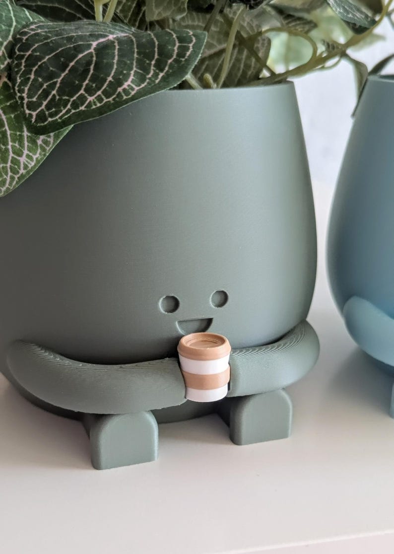 May include: A green ceramic planter shaped like a person with a smiling face and arms holding a small brown and white striped cup. The planter has a leafy green plant growing out of the top.