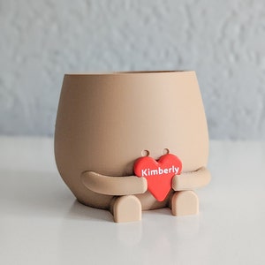 Puede incluir: Un macetero de cer&aacute;mica marr&oacute;n con forma de persona que sostiene un coraz&oacute;n rojo con el nombre Kimberly.