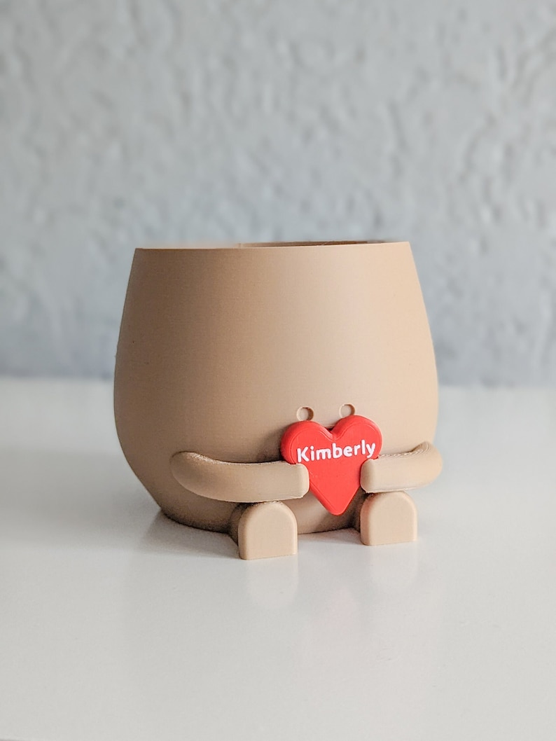 Puede incluir: Un macetero de cer&aacute;mica marr&oacute;n con forma de persona que sostiene un coraz&oacute;n rojo con el nombre Kimberly.