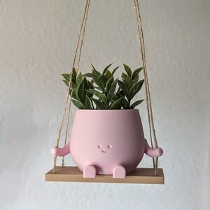 K&ouml;nnte beinhalten: Ein rosa Blumentopf mit l&auml;chelndem Gesicht, Armen und Beinen, gef&uuml;llt mit gr&uuml;nen Pflanzen. Der Topf h&auml;ngt an einem Seil und ruht auf einem kleinen Holzregal. Ein dekorativer Artikel f&uuml;r Zimmerpflanzen.