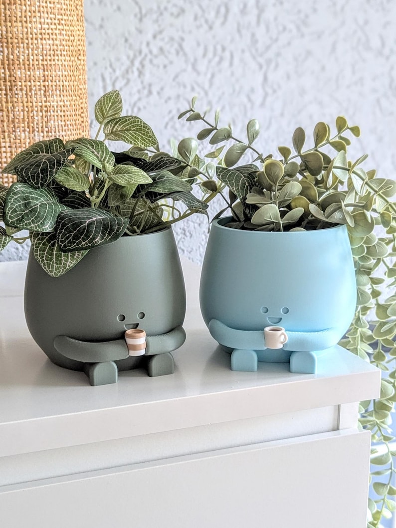 May include: Two ceramic plant pots with cartoon faces and arms holding small cups. The pot on the left is dark green and the pot on the right is light blue. Both pots have plants growing out of them.