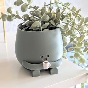 May include: A green ceramic planter shaped like a cartoon character holding a small white cup. The planter has a happy face and is holding a cup of coffee. The planter is filled with a green leafy plant.