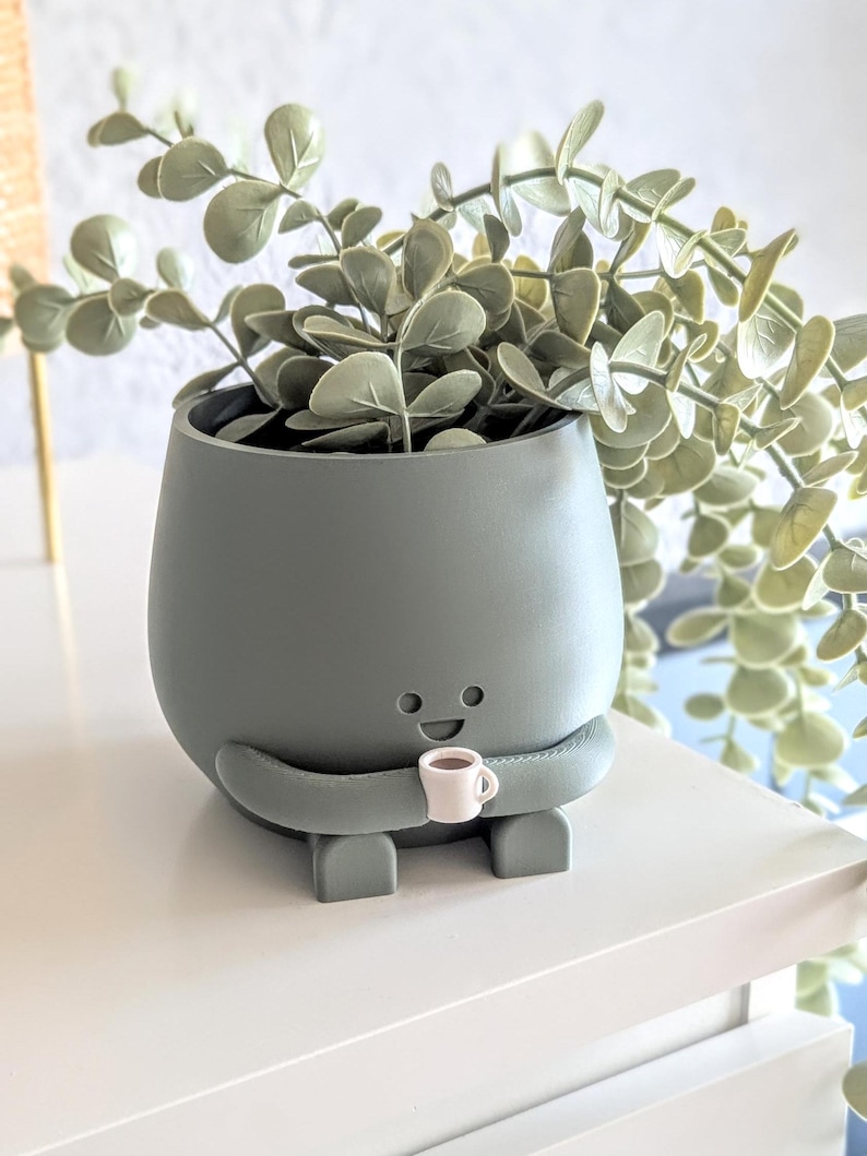 May include: A green ceramic planter shaped like a cartoon character holding a small white cup. The planter has a happy face and is holding a cup of coffee. The planter is filled with a green leafy plant.