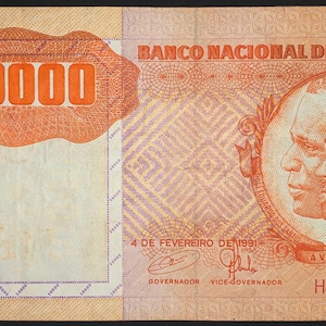 Pode incluir: Nota de 100.000 Kwanzas de Angola. A nota apresenta o texto "BANCO NACIONAL DE ANGOLA" e "100000". A nota tem um retrato de dois homens e o texto "A VITORIA E CERTA". A nota é principalmente laranja, azul e verde.