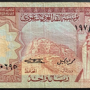 May include: A one Saudi riyal banknote featuring a portrait of King Faisal on the right side, an illustration of a mountain landscape in the center, and intricate geometric patterns in shades of brown, tan, and cream with Arabic text throughout.