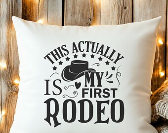 May include: White square pillow with the text "THIS ACTUALLY IS MY FIRST RODEO" in black. The design includes a cowboy hat, stars, and decorative swirls. The pillow is on a wooden surface.