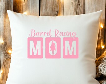 May include: White square pillow with the text "Barrel Racing MOM" in pink. The word "MOM" is in large pink blocks, with a silhouette of a rider on a horse in the center. The pillow is on a wooden surface.