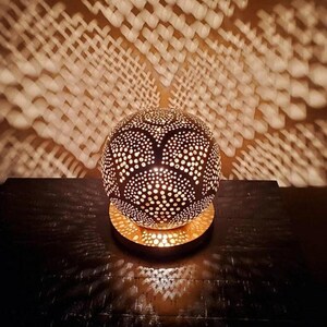 Puede incluir: Una lámpara redonda de metal con un diseño decorativo recortado. La lámpara está encendida y proyecta una sombra con patrón en la superficie de abajo.