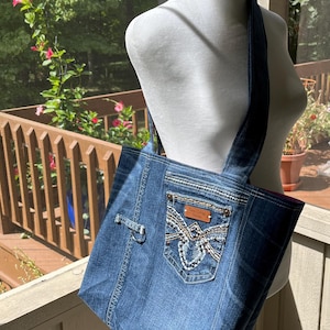 May include: A blue denim tote bag with a large pocket on the front. The pocket has a brown leather patch with the text "Rock & Roll Denim" on it. The bag has a brown leather strap and a silver buckle.