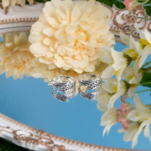 May include: Two silver rings with intricate designs, one engraved with "Andrea" and the other with "Jason", reflected in a blue mirrored surface.
