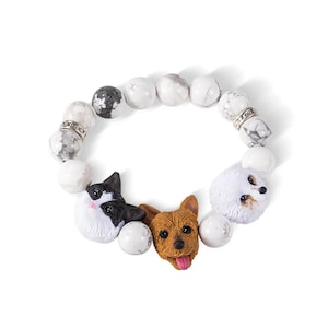 May include: White stone bead bracelet with three charms: a black and white cat, a brown dog, and a white dog. The bracelet is made of white stones with a silver accent.