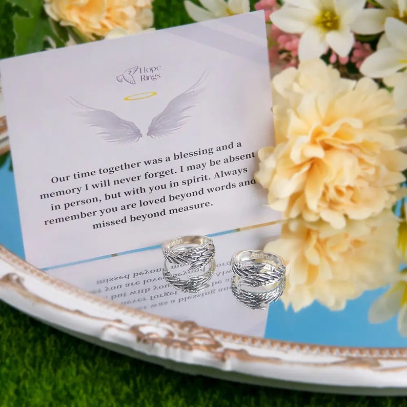 May include: Two silver rings with a wing design. The rings are on a white surface with a card that reads "Hope Rings" and a message about remembering a loved one. The card is surrounded by white and yellow flowers.