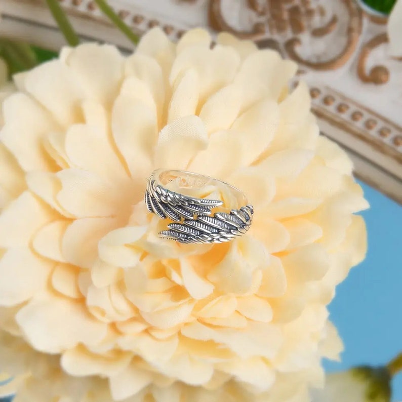 May include: Silver ring with a detailed design of angel wings. The ring is on a white flower.
