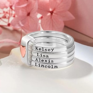 May include: A silver ring with four bands, each engraved with a name: Kelsey, Lisa, Alexis, and Lincoln. A small heart-shaped accent in rose gold is on the side. The ring is on a white surface with a soft pink floral background.