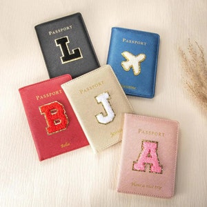 Könnte beinhalten: Fünf Reisepasshüllen mit verschiedenen Farben und personalisierten Initialen oder Designs. Die Hüllen sind schwarz, blau, rot, beige und rosa. Die Initialen sind L, X, B, J und A. Der Text auf den Hüllen lautet "Passport" und "Have a nice trip".