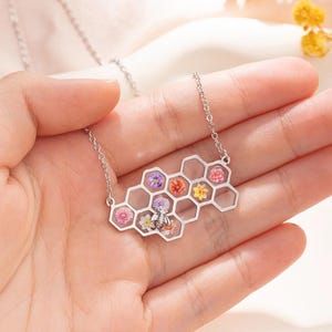 May include: A silver chain necklace with a honeycomb pendant featuring a bee and pressed flowers in various colours.